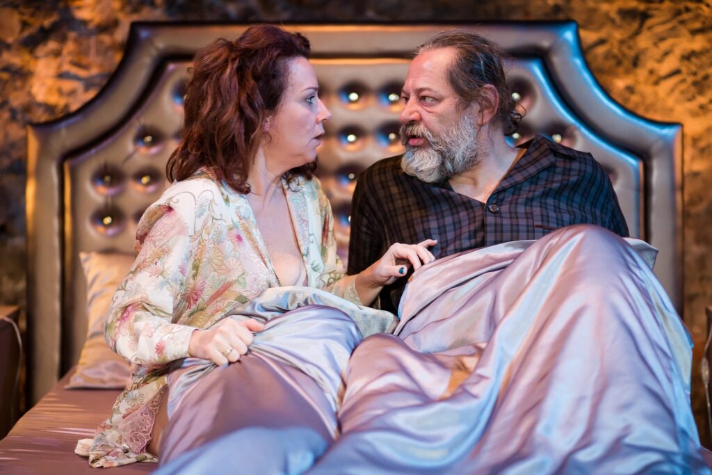 Zdjęcie ze spektaklu Via-gra. Zbliżenie. Aktorka w szlafroku i aktor z długą siwą brodą w piżamie siedzą na łóżku. Są przykryci kołdrą i patrzą na siebie.