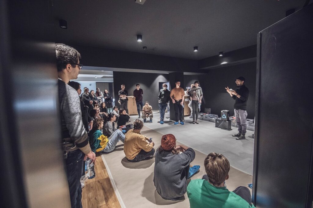 Zdjęcie z realizacji projektu PLAYON. Duża grupa ludzi, część osób siedzi na podłodze, Skierowani są w stronę lidera, który przemawia.