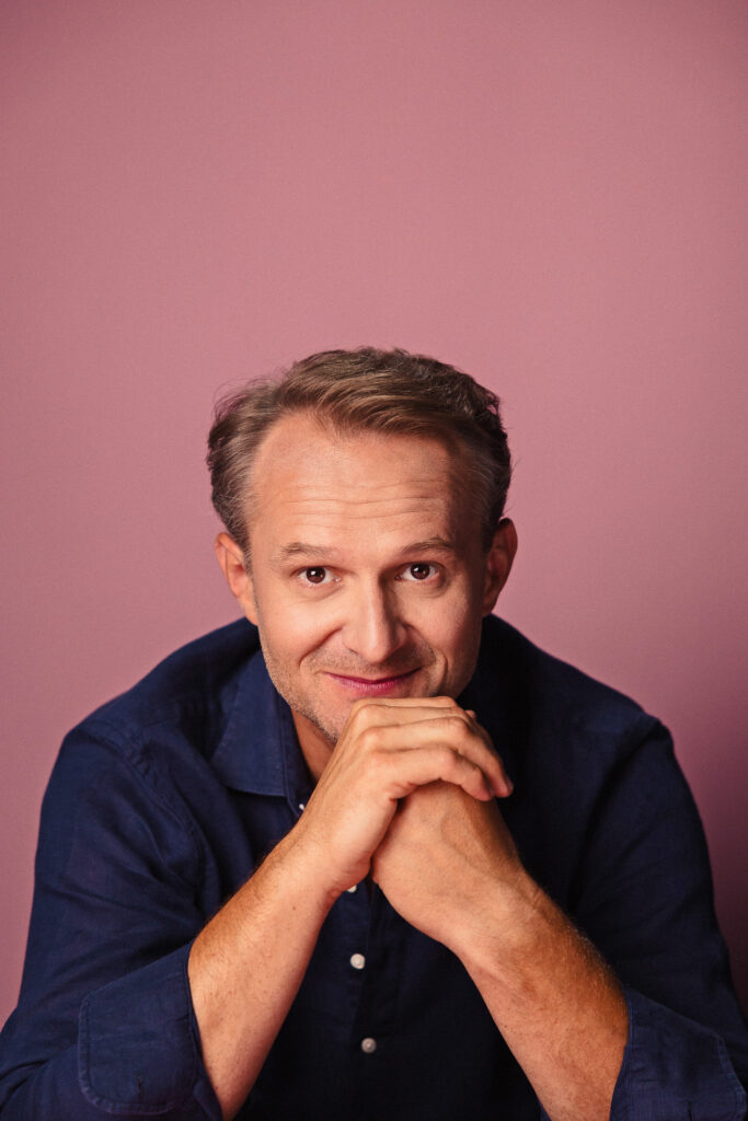 Zdjęcie półprofil portretowe aktora Pawła Kumięgi, lekki uśmiech, bliski kadr, ręce splecione przed twarzą.