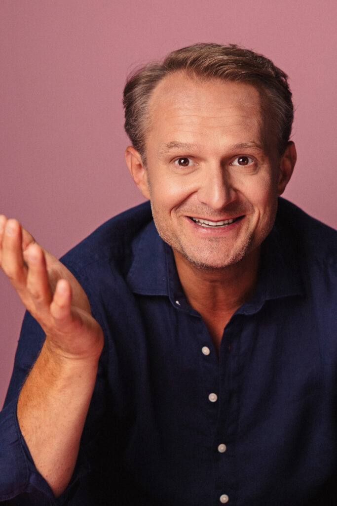 Zdjęcie półprofil portretowe aktora Pawła Kumięgi, szeroki uśmiech, bliski kadr, ręka wyciągnięta do obiektywu.