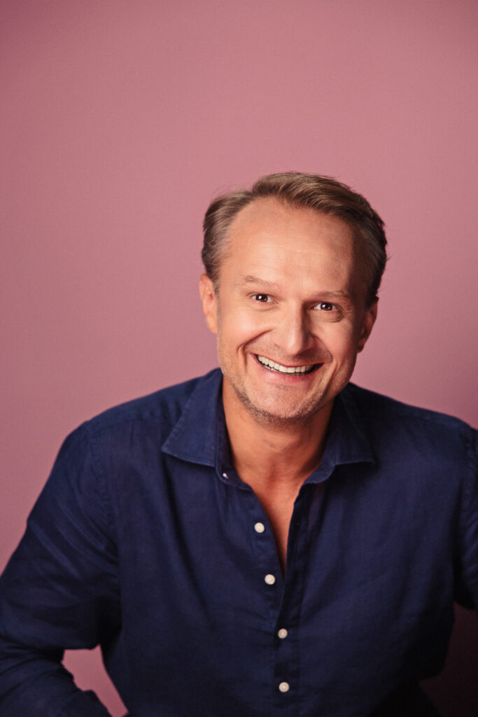 Zdjęcie portretowe aktora Pawła Kumięgi, szeroki uśmiech, bliski kadr.