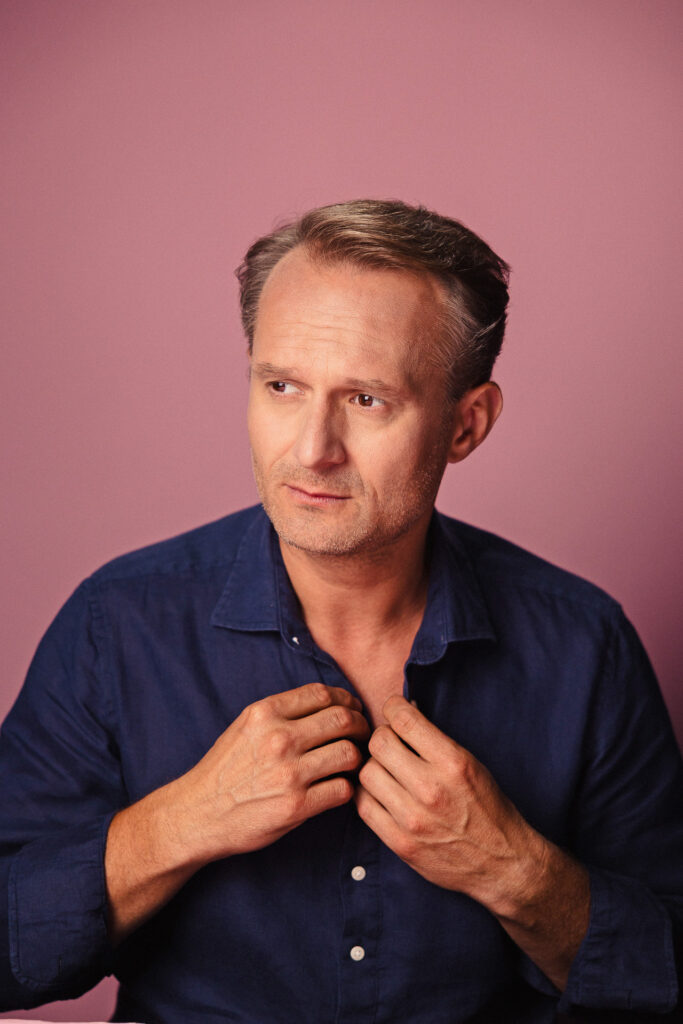 Zdjęcie półprofil portretowe aktora Pawła Kumięgi, poważna twarz, ręce przy koszuli kołnierzyku.