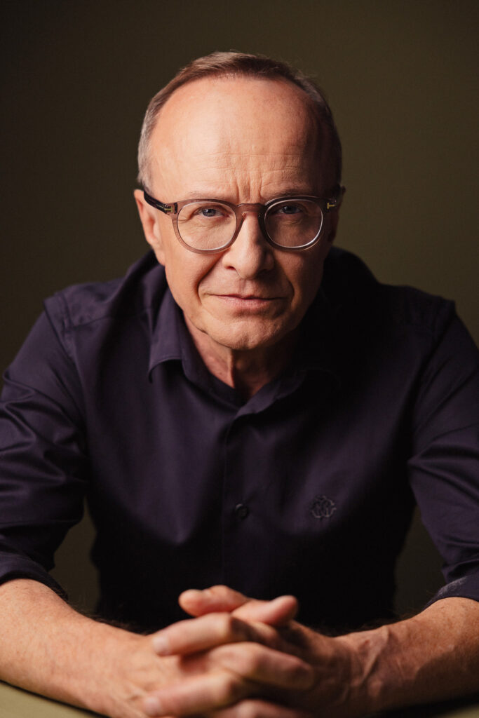 Zdjęcie portretowe aktora Piotra Pilitowskiego, lekki uśmiech, ręce skrzyżowane przed sobą.