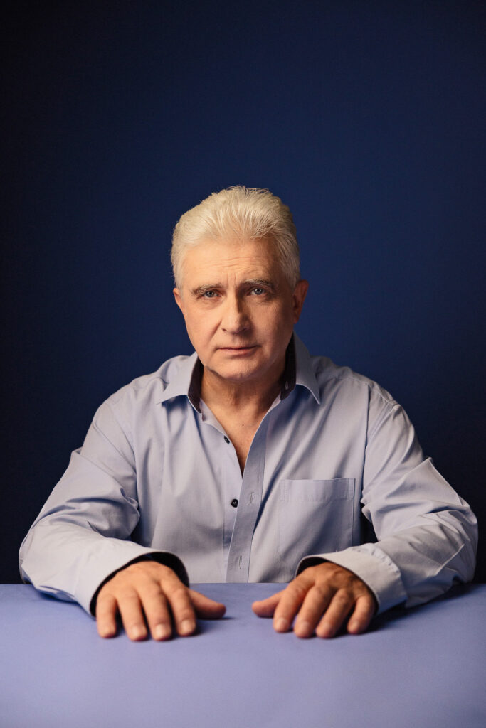 Zdjęcie portretowe aktora Piotra Piechy, poważna twarz, ręce na wprost sylwetki.