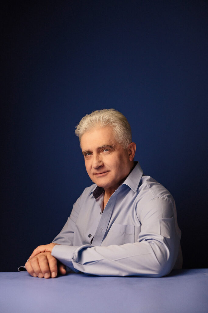 Zdjęcie portretowe aktora Piotra Piechy, poważna twarz, ręce splecione przed sobą.