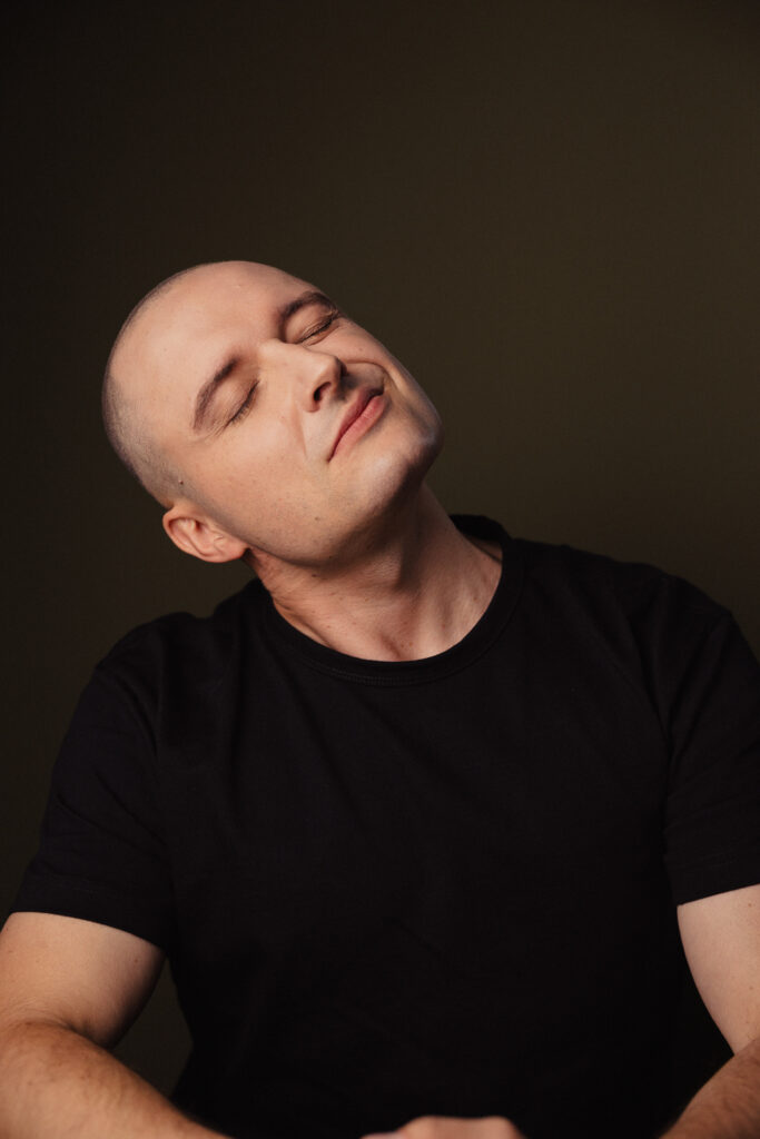Zdjęcie portretowe aktora Patryka Palusińskiego, odprężona twarz, przechylona głowa zamknięte oczy, bliski kadr.