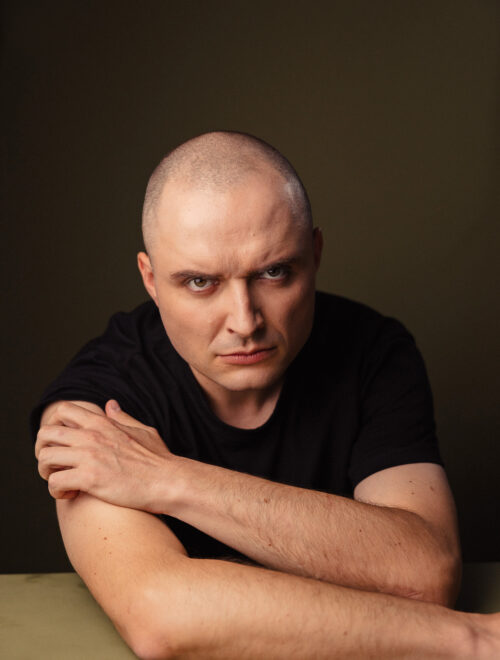 Zdjęcie portretowe aktora Patryka Palusińskiego, poważna twarz, ręce skrzyżowane przed sobą.