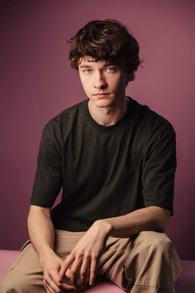 Zdjęcie portretowe aktora Marcina Mazurka, poważna twarz, daleki kadr, ręce spoczywają na nogach.