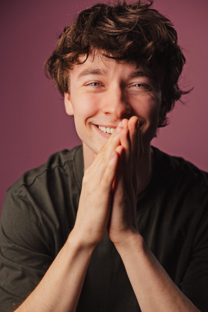 Zdjęcie portretowe aktora Marcina Mazurka, szeroki uśmiech, bliski kadr, ręce częściowo zasłaniają twarz złożone przed sobą.