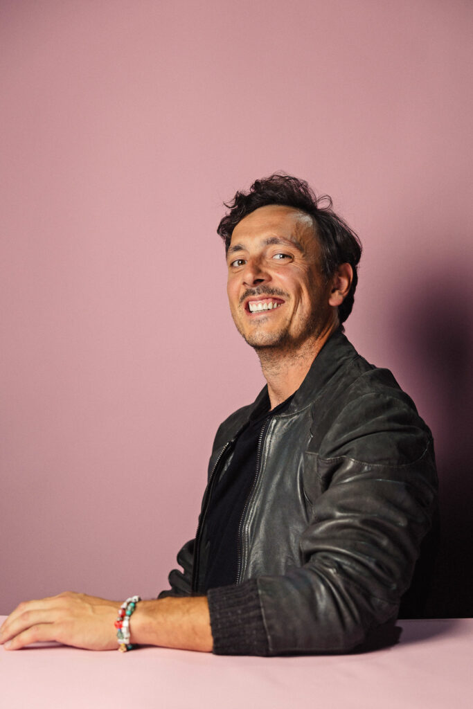 Zdjęcie półprofil profilowe portretowe aktora Piotra Franasowicza, szeroki uśmiech, ręce przed sobą.