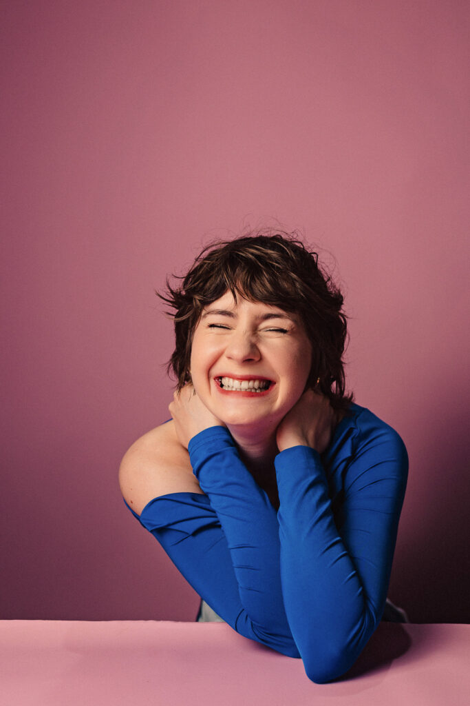 Zdjęcie portretowe aktorki Marii Chojnackiej, szeroki uśmiech, ręce blisko sylwetki dotykają szyi.