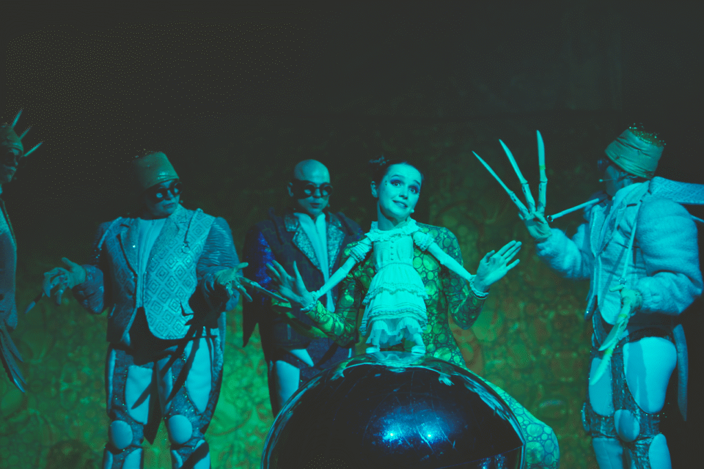 Zdjęcie ze spektaklu Calineczka. Scena zbiorowa, zdjęcie zabarwione na zielono. Aktorka z przyczepioną do kostiumu lalką, siedzi na dużej szklistej kuli. Wokół niej blisko stoją aktorzy przebrani za chrabąszcze w połyskujących kostiumach.