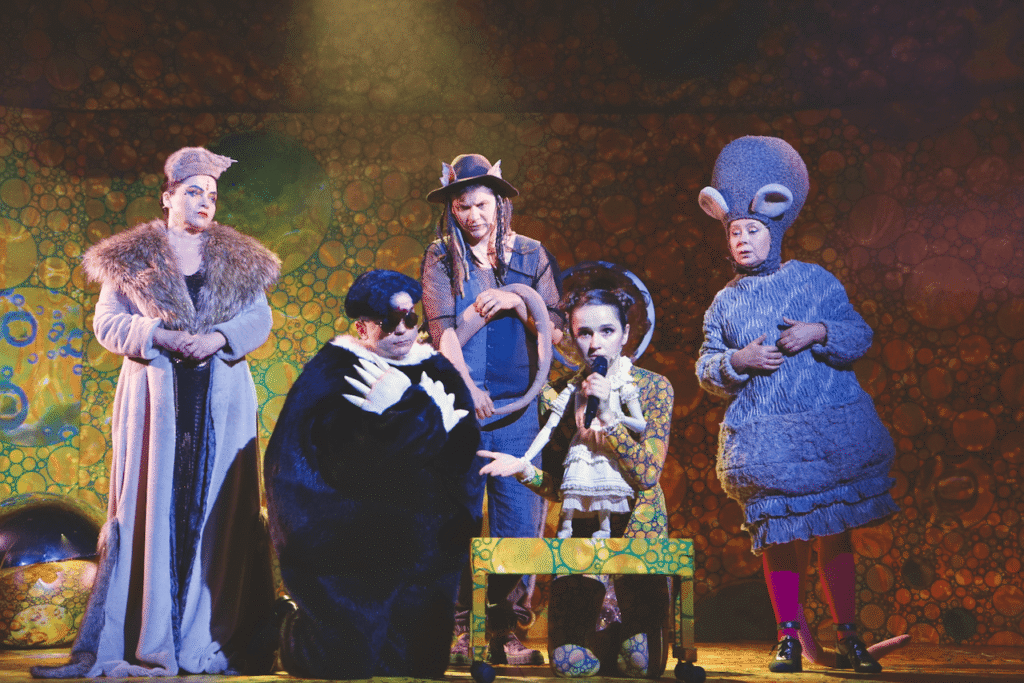 Zdjęcie ze spektaklu Calineczka. Aktorka z przyczepioną do kostiumu lalką, siedzi na ławeczce. Obok niej kuca aktor w czarnym kostiumie, za nim stoi aktorka w futrze oraz aktor w czapce z uszami. Z prawej strony stoi aktorka przebrana za mysz.