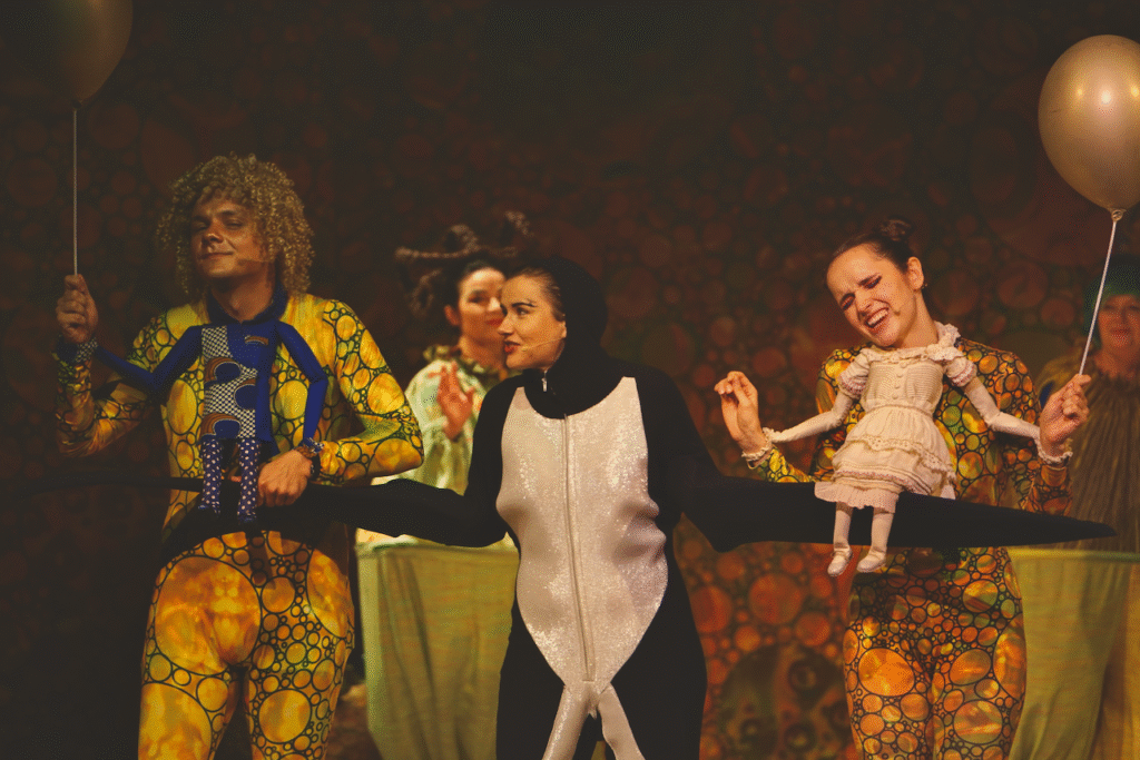 Zdjęcie ze spektaklu Calineczka. Scena zbiorowa. Aktorka z przyczepioną do kostiumu lalką- Calineczka, siedzi na ręce aktorki przebranej za jaskółkę, po drugiej stronie stoi aktor z przyczepioną lalką chłopca - ma złote włosy i twarz.