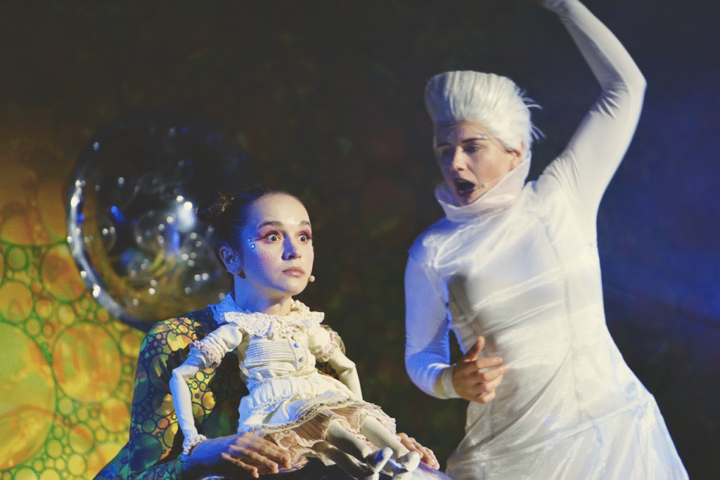 Zdjęcie ze spektaklu Calineczka. Aktorka z przyczepioną do kostiumu lalką, siedzi na dużej szkliste kuli. Ma zaskoczoną minę, obok niej stoi aktorka w białym kostiumie i białej peruce, wykonuje ruch ręką - czaruje.