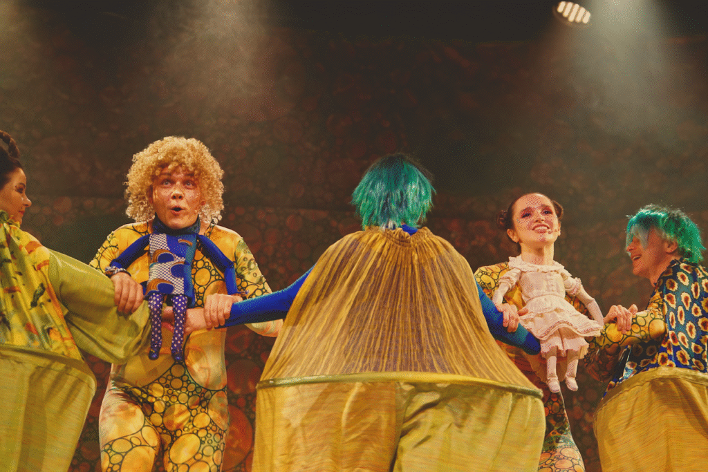 Zdjęcie ze spektaklu Calineczka. Scena zbiorowa.Aktorka z przyczepioną do kostiumu lalką, siedzi na ręce aktorki przebranej za elfa - duży obszary bufiasty kostium, po drugiej stronie stoi aktor z przyczepioną lalką chłopca - ma złote włosy i twarz.