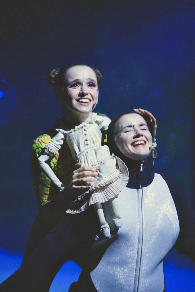 Zdjęcie ze spektaklu Calineczka. Aktorka z przyczepioną do kostiumu lalką - Calineczka. Ma uśmiechniętą twarz, siedzi na ramieniu drugiej aktorki przebranej za jaskółkę, biało czarny kombinezon. Jest uśmiechnięta, śpiewa.