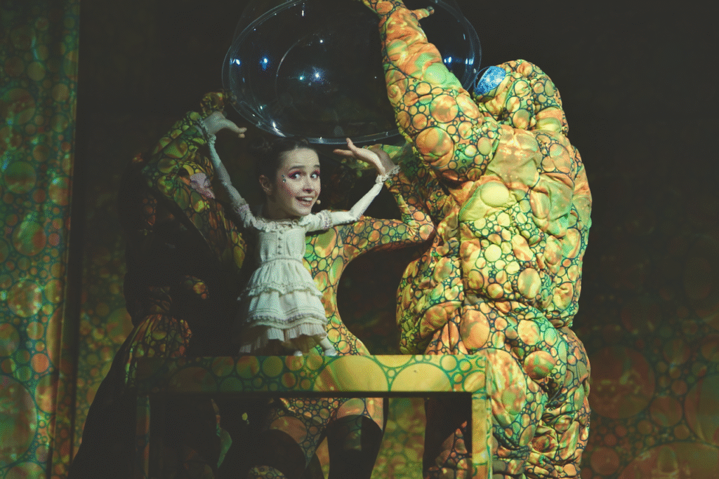 Zdjęcie ze spektaklu Calineczka. Scena zbiorowa.Aktorka z przyczepioną do kostiumu lalką- Calineczka, siedzi na ławeczce. Obok niej dwoje aktorów przebranych za żaby próbują nałożyć na nią przeszkloną kulę.