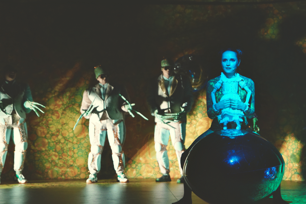 Zdjęcie ze spektaklu Calineczka. Scena zbiorowa. Aktorka z przyczepioną do kostiumu lalką, siedzi na dużej szkliste kuli jest oświetlona na niebiesko. Za nią stoją aktorzy przebrani za chrabąszcze, na rękach mają szczypce, ich kostiumy połyskują.