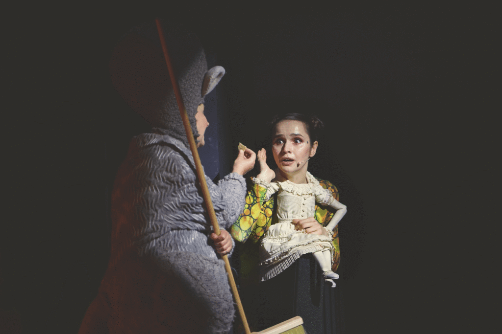 Zdjęcie ze spektaklu Calineczka. Aktorka z przyczepioną do kostiumu lalką, siedzi na dużej szkliste kuli. Ma przestraszoną minę. Obok niej plecami stoi aktorka przebrana za mysz, podaje ciastko pierwszej aktorce, w drugiej trzyma miotłę.