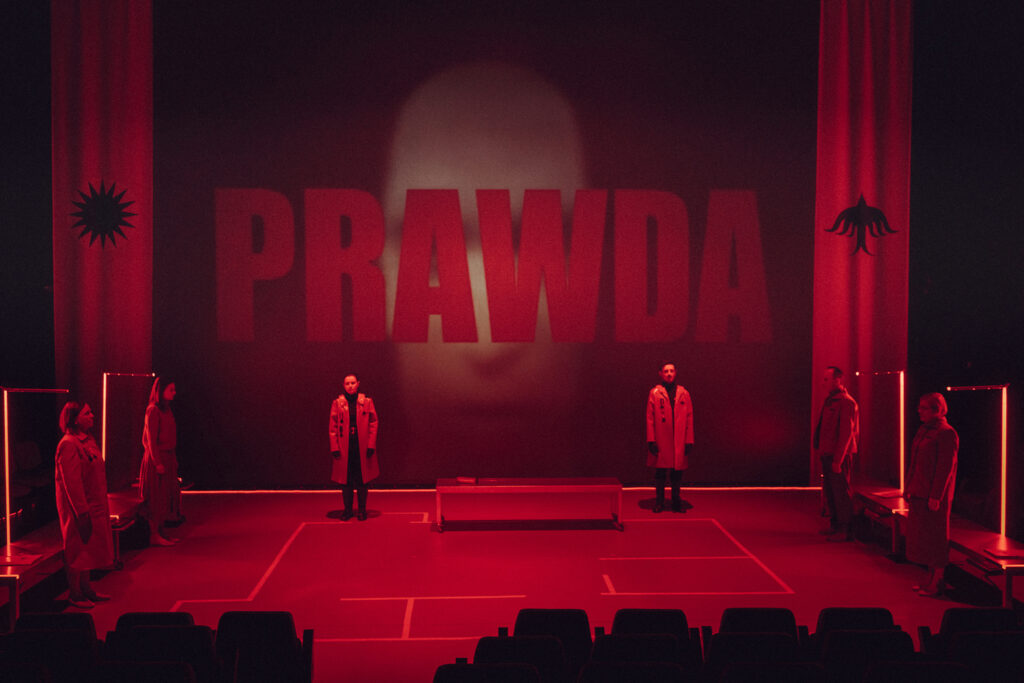 Zdjęcie ze spektaklu Ministerstwo miłości. W oddali stoją dwie postaci, za ich plecami na dużym ekranie wyświetla się słowo prawda, zdjęcie zabarwione na czerwono.