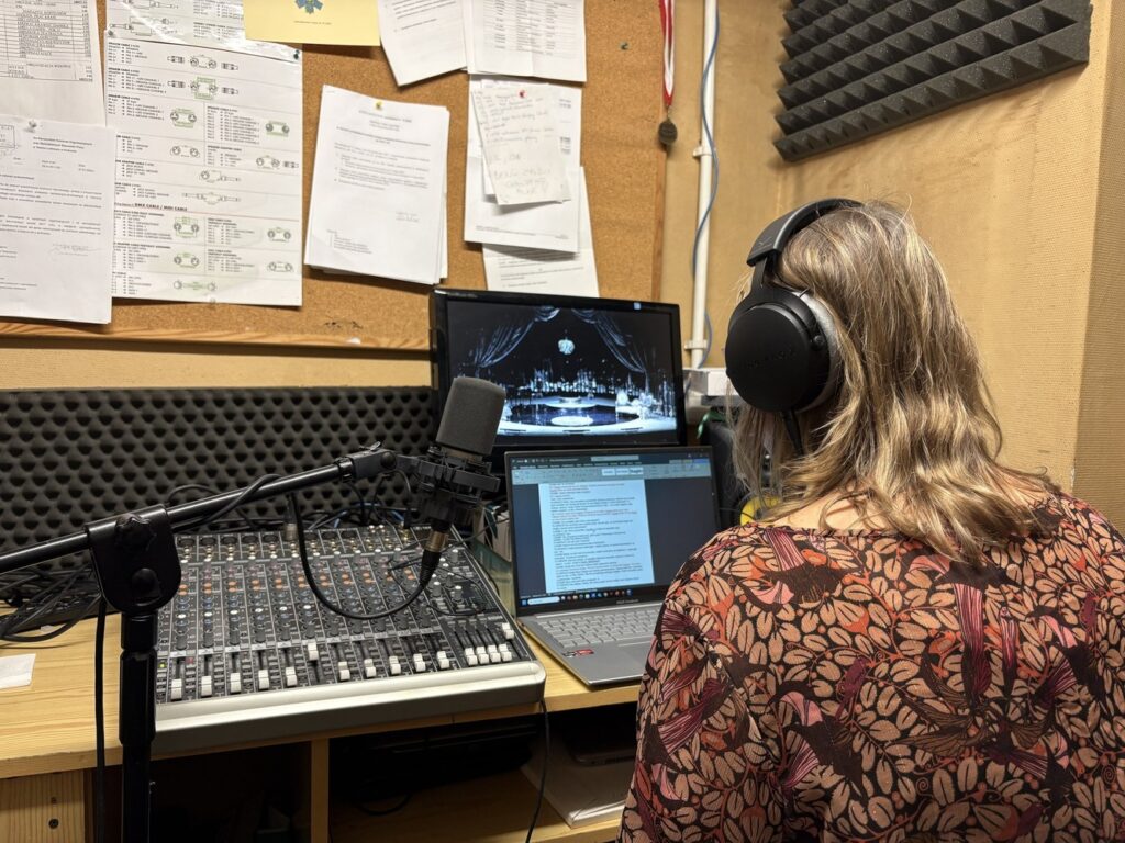 Tyłem siedzi kobieta w słuchawkach, siedzi przed monitorem i pulpitem akustycznym obsługuje audiodeskrypcję.