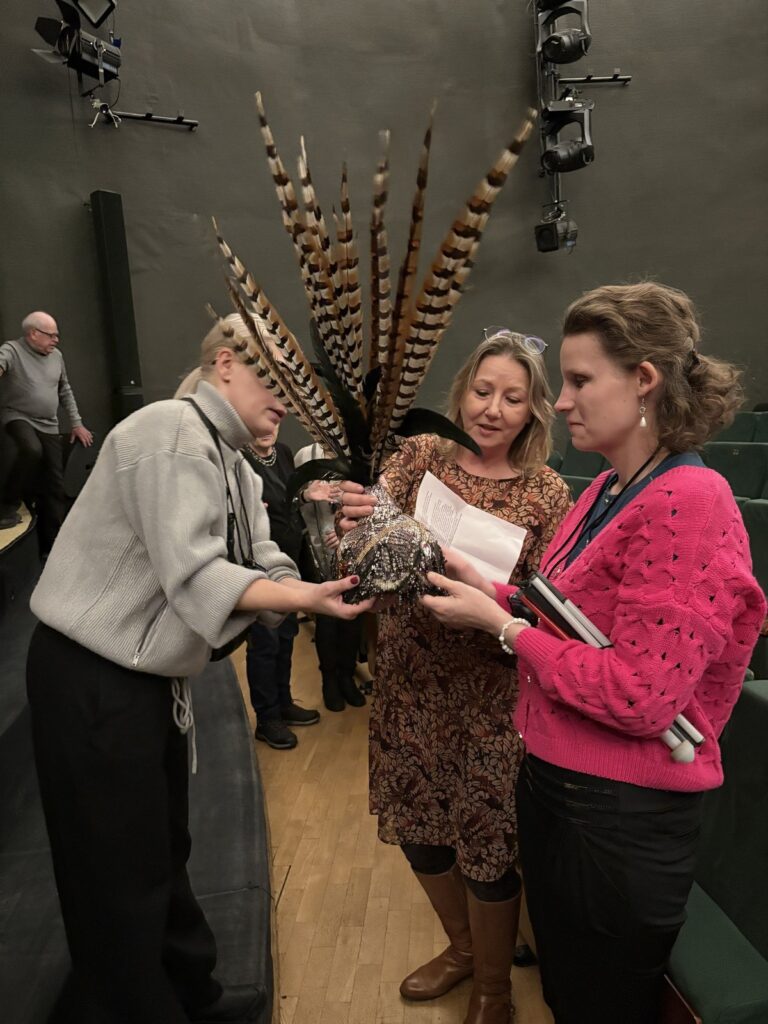 Trzy kobiety, jedna obserwuje jak dwie oglądają kapelusz z długimi piórami i koralikami.