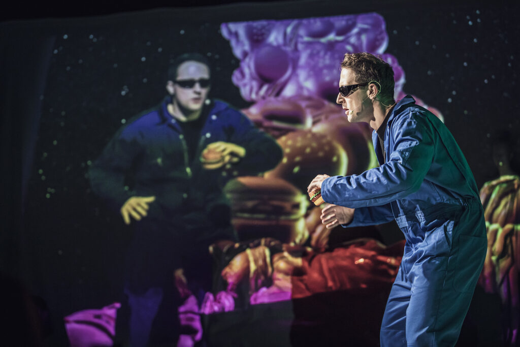 Zdjęcie ze spektaklu Co gryzie Gilberta Grape'a. Z prawej strony aktor w niebieskim kombinezonie w okularach i z hamburgerem w ręku po prawej stronie widzimy go na ekranie.