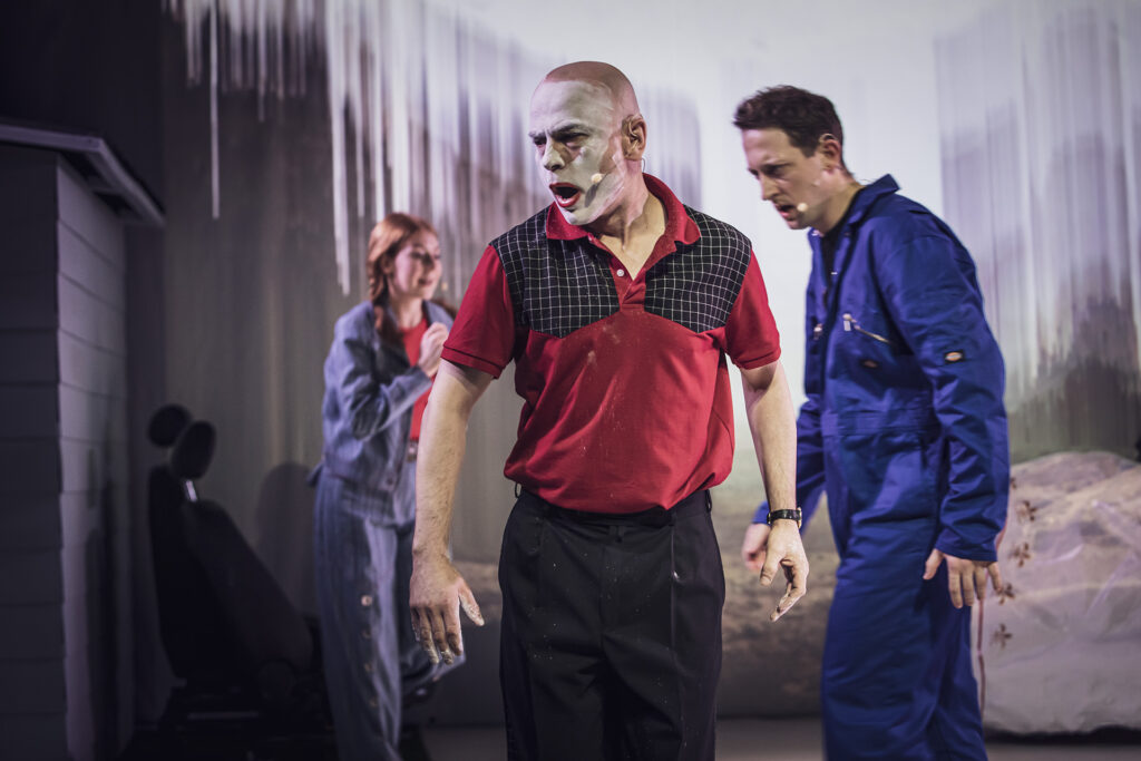 Zdjęcie ze spektaklu Co gryzie Gilberta Grape'a. Na pierwszym planie aktor z wymalowaną twarzą na biało w zastygniętej pozie, w tle rozmazane sylwetki dwóch osób.