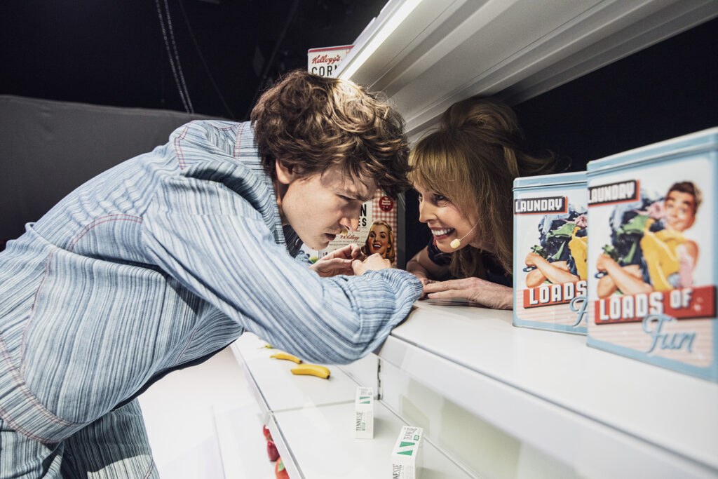 Zdjęcie ze spektaklu Co gryzie Gilberta Grape'a. Dwie osoby opierają się o półki sklepowe spoglądają na siebie wokół nich są towary.