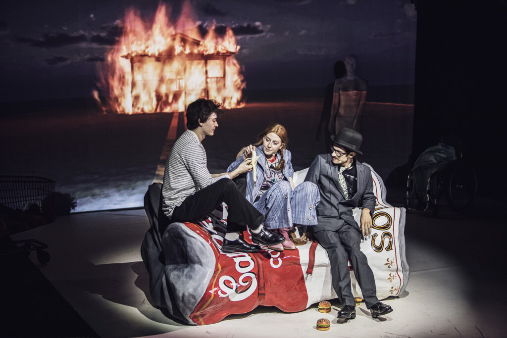 Zdjęcie ze spektaklu Co gryzie Gilberta Grape'a. Troje młodych aktorów siedzi na kanapie, która wygląda jak zgnieciona puszka po zupie. W tle widzimy płonący dom na ekranie.