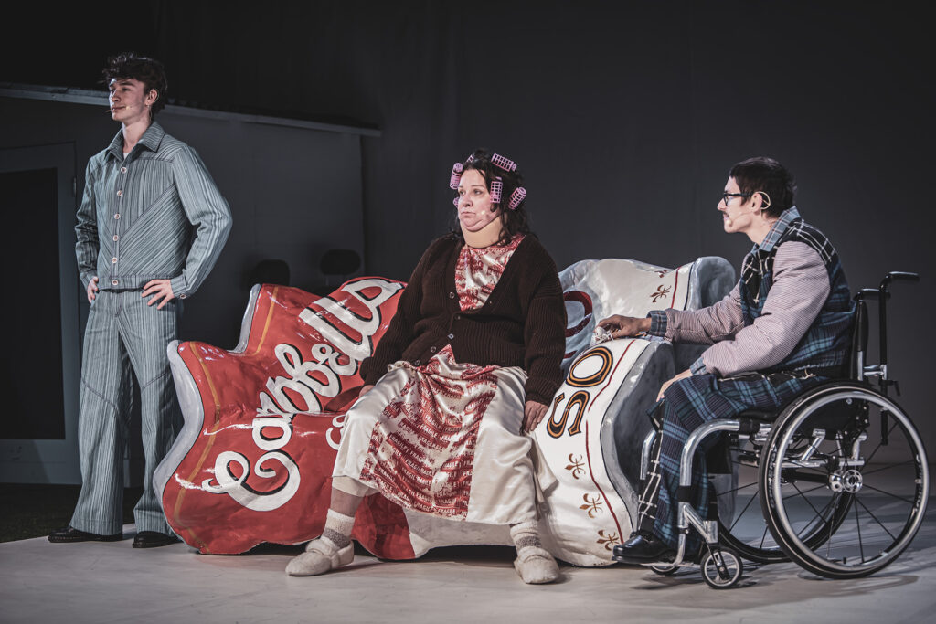 Zdjęcie ze spektaklu Co gryzie Gilberta Grape'a. Młody aktor stoi sztywno z lewej strony na kanapie siedzi kobieta z wałkami na głowie, z prawej strony młody aktor na wózku.