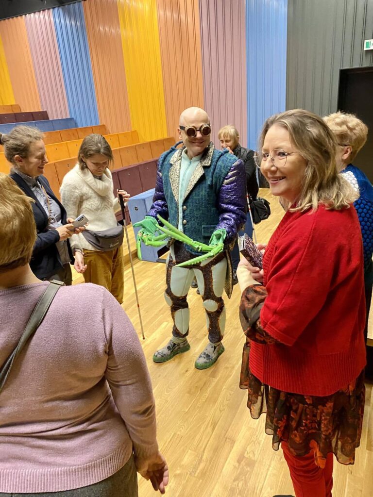 Grupa ludzi stoi w okół aktora w błyszczącym kostiumie chrząszcza, ma rękawiczki przypominające duże szczypce.