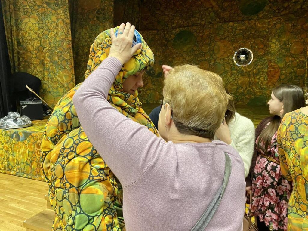Kobieta stoi tyłem, ma wyciągnięte ręce dotyka głowy aktora w stroju żaby.