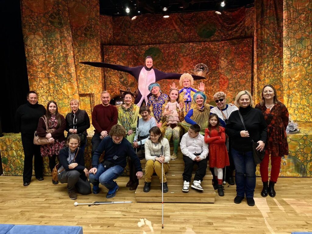 Zdjęcie grupowe, aktorzy w kostiumach i osoby z niepełnosprawnością wzroku i ich osoby towarzyszone pozują do zdjęcia przed sceną