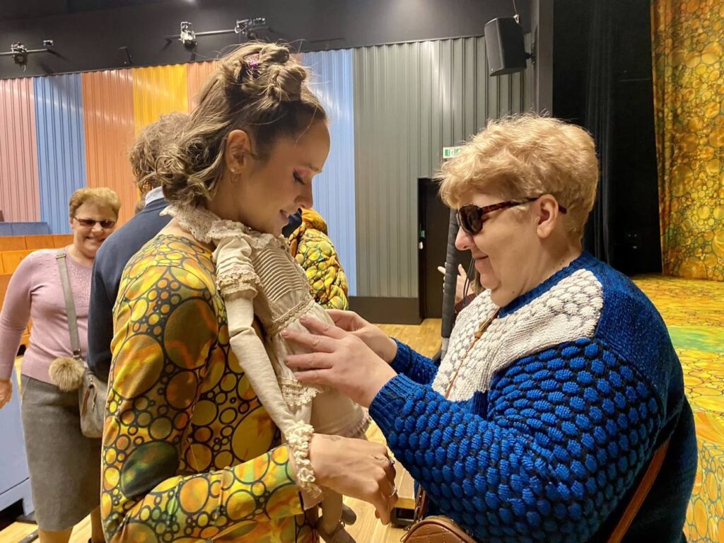 Dwie kobiety. Aktorka w kostiumie Calineczki prezentuje drugiej kobiecie lalkę Calineczki, którą ma przyczepioną do szyi.