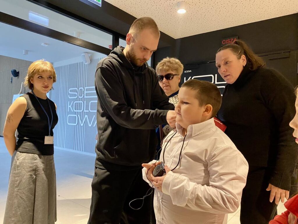 Na teatralnym korytarzu stoi grupa ludzi , mężczyzna pomaga załorzyć słuchawkę do audiodeskrypcji małemu chłopcu