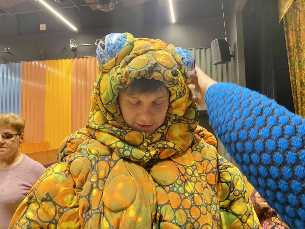 Zbliżenie na aktora w kostiumie żaby. Kolorowy kostium na głowiem duże oczy, w ich kierunku jest wyciągnięta ręka.