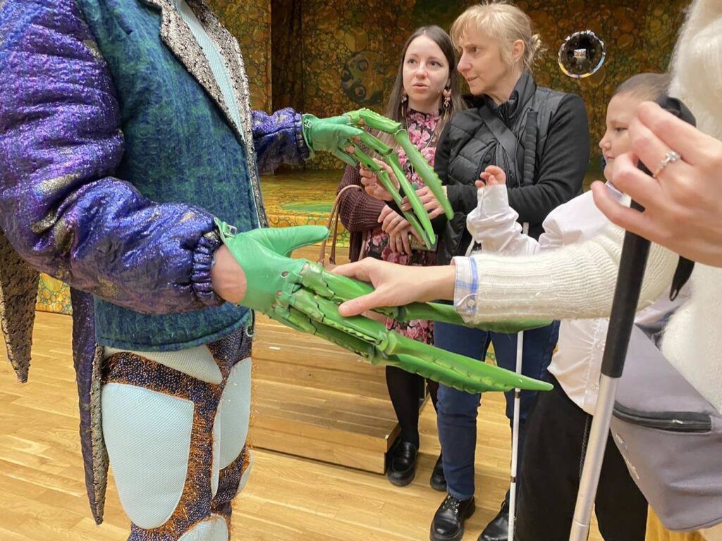 Zbliżenie na dłonie aktora, na których są rękawiczki przypominające szczypce chrząszcz. Osoba przed nim dotyka je.