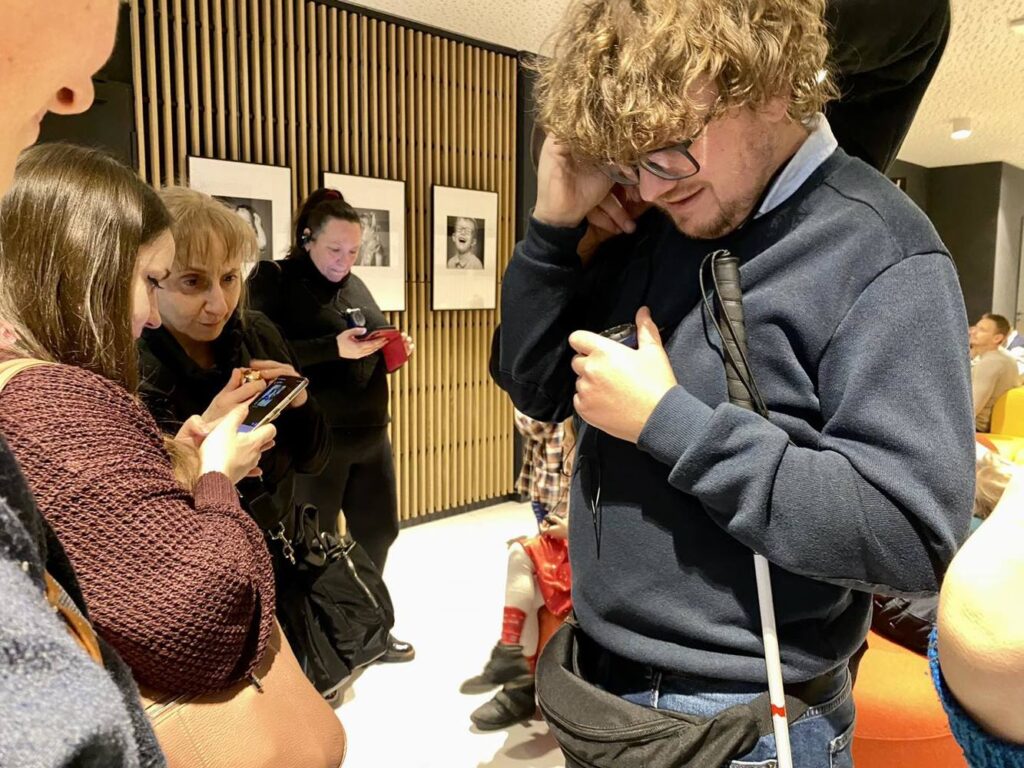 Na korytarzu teatralnym stoi grupa ludzi, mężczyzna na pierwszym planie zakłada słuchawkę do audiodeskrypcji.