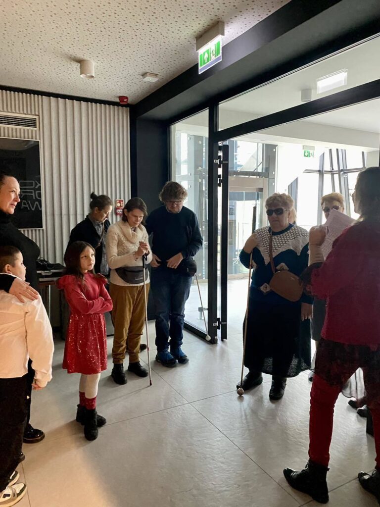 Zdjęcie grupowe. Grupa ludzi stoi i słucha audiowbtępu, który mówi lektorka przed spektaklem.