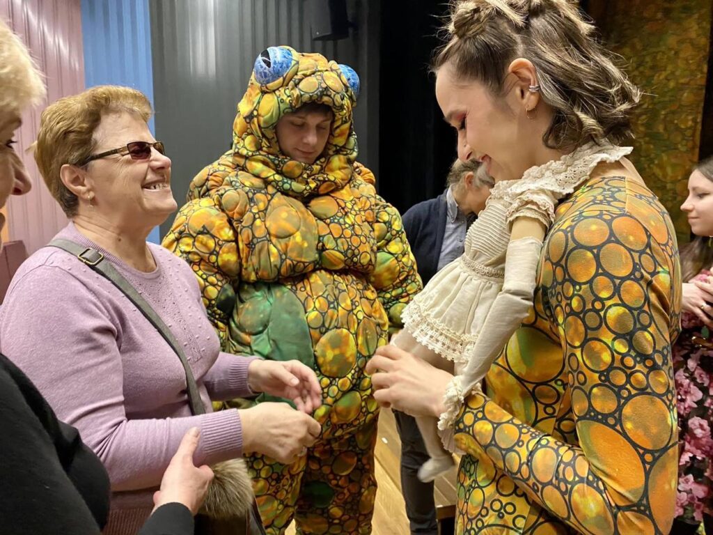 Osoba z niepełnosprawnością wzroku stoi przed aktorką grającą Calineczkę. Aktorka ma do szyi przyczepioną lalkę/ ciało Calineczki, w tle aktor w stroju żaby.