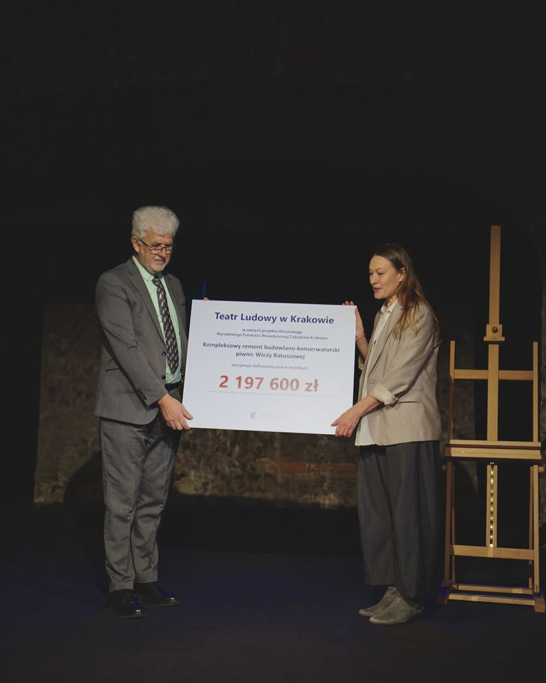 Na Scenie stoją dwie osoby które trzymają w rękach tablicę z promesą na dofinansowanie kompleksowego remontu budowlano - konserwatorskiego piwnic Wieży Ratuszowej