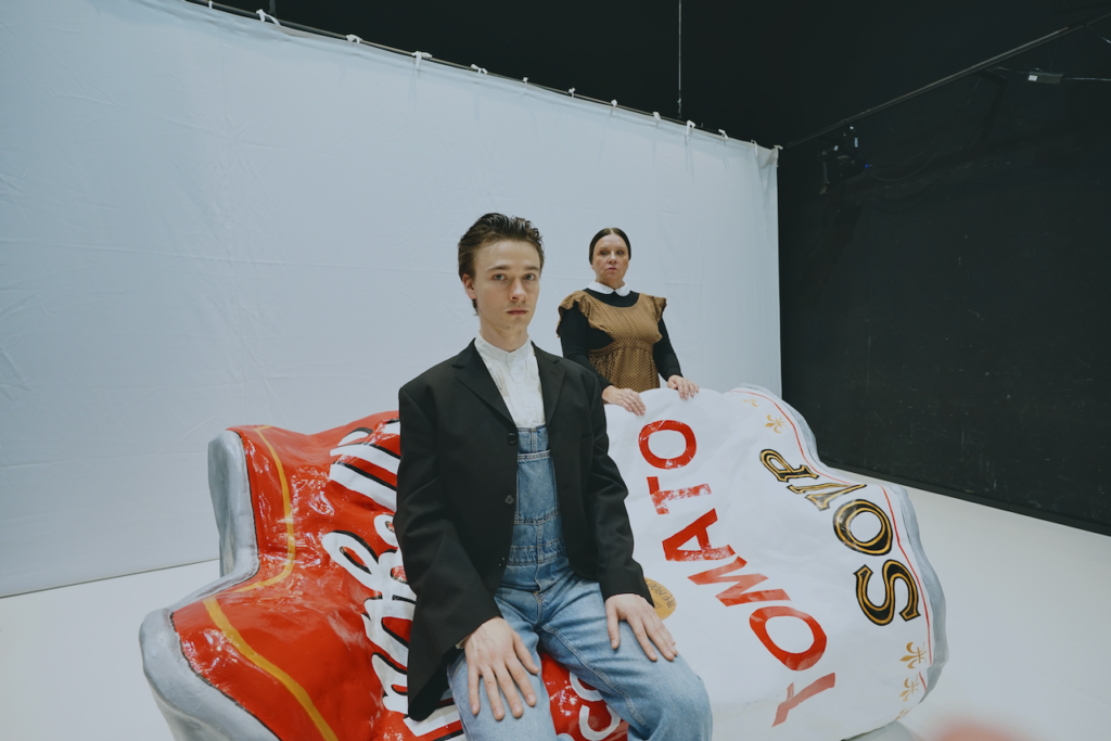 Pozowana sytuacja. Aktor w ogrodniczkach i marynarce siedzi na kanapie przypominającej zgniecioną puszkę, za jego plecami stoi aktorka w brązowym fartuchu. W tle biały ekran.