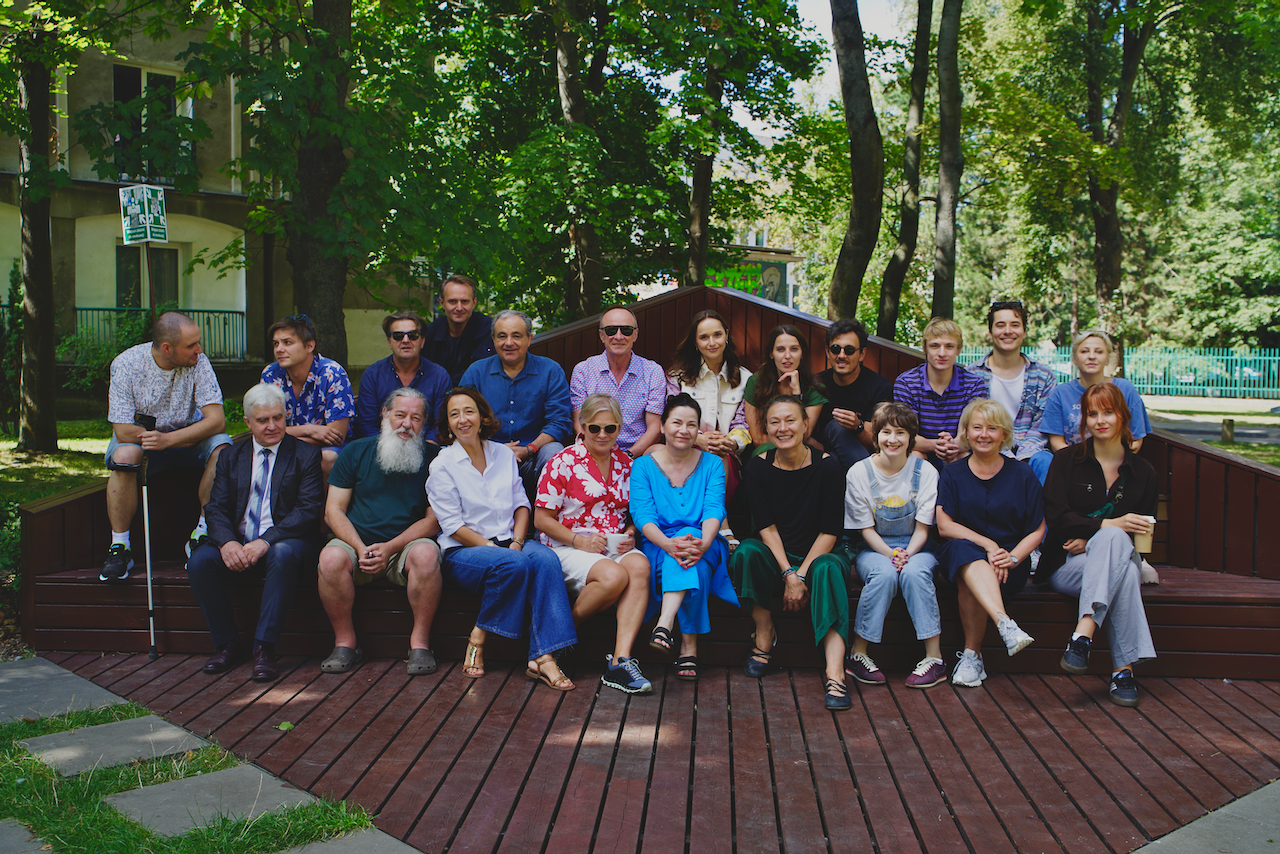 Zdjęcie pozowane, grupowe. Na zewnątrz w słoneczną pogodę na dużej ławce siedzą aktorzy i aktorki oraz twórcy spektaklu Uroczystość.