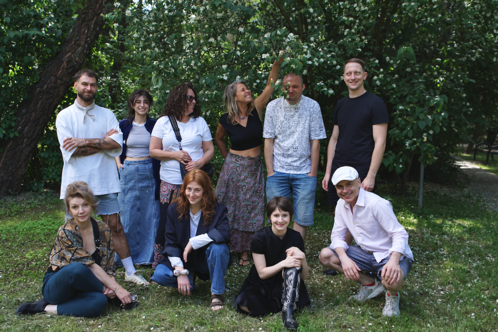 Grupa ludzi pozuje do zdjęcia na tle zieleni, Cześć osób stoi, część kuca. Są weseli, uśmiechają się.