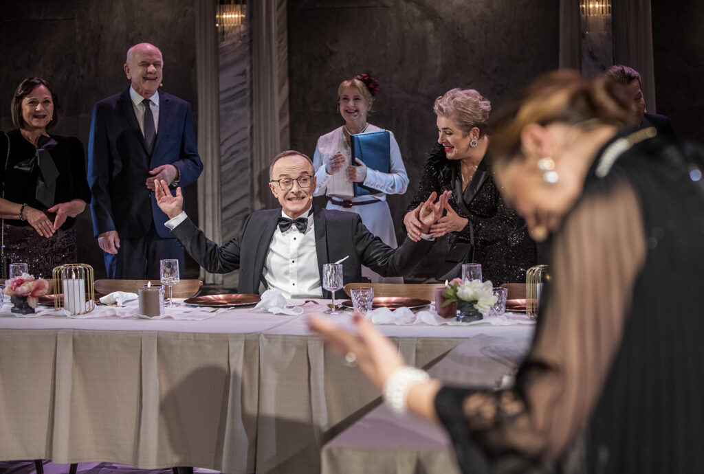 Zdjęcie ze spektaklu Uroczystość. Zdjęcie grupowe, za stołem z otwartymi ramionami siedzi mężczyzna, w okół niego roześmiani ludzie.