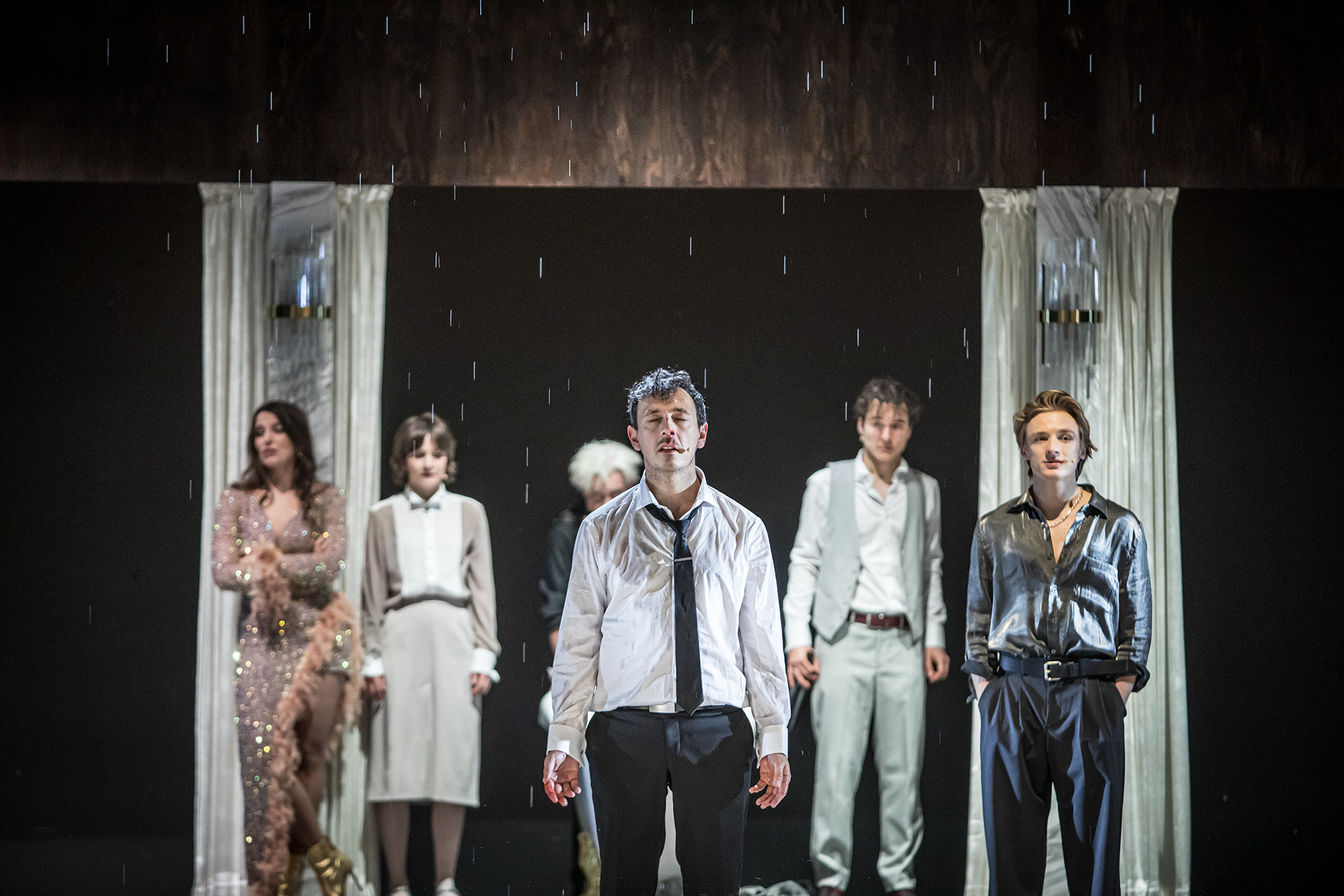 Zdjęcie ze spektaklu Uroczystość. Po środku stoi mężczyzna w zmoczonym ubraniu, widać krople wody spadające z sufitu. W tle grupa ludzi.