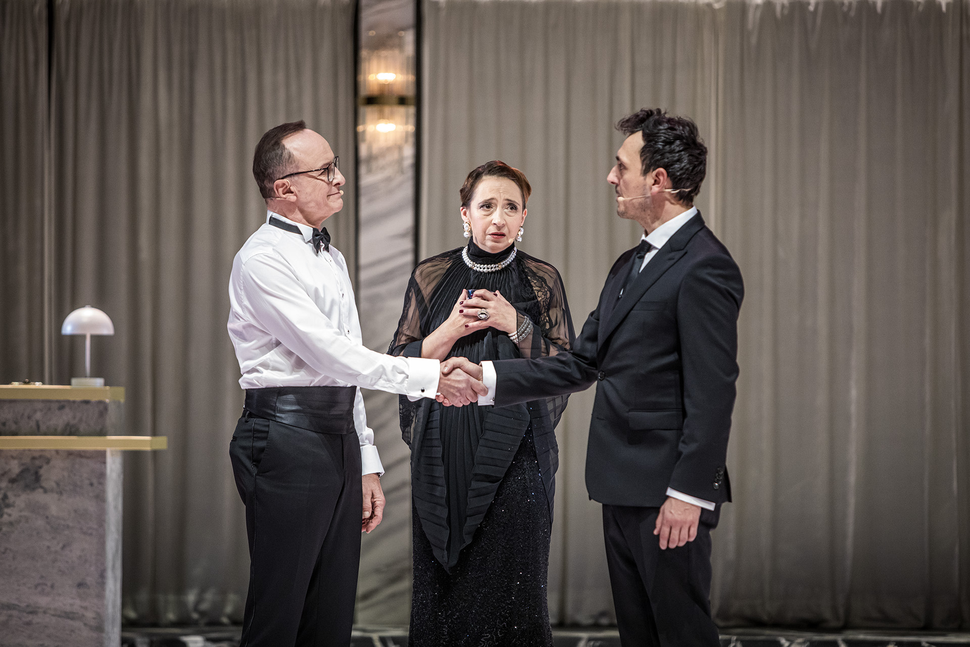 Zdjęcie ze spektaklu Uroczystość. Troje elegancko ubranych aktorów. Dwóch mężczyzn podaje sobie ręce, kobieta ma zaniepokojoną minę.