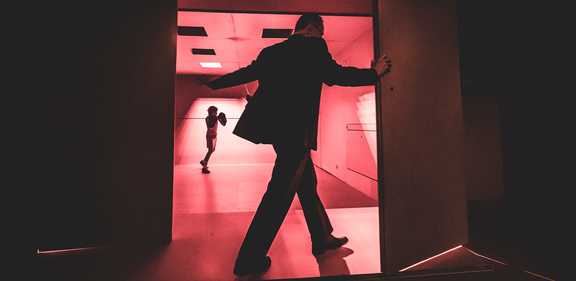 Śmierć komiwojażera. zdjęcie ze spektaklu. Czerwona scena i ściany, aktor otwiera drzwi , za drzwiamy mały chłopiec bijący worek treningowy